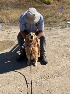 Golden retriever Cali sits in front of Sid, in a blue shirt and tan hat