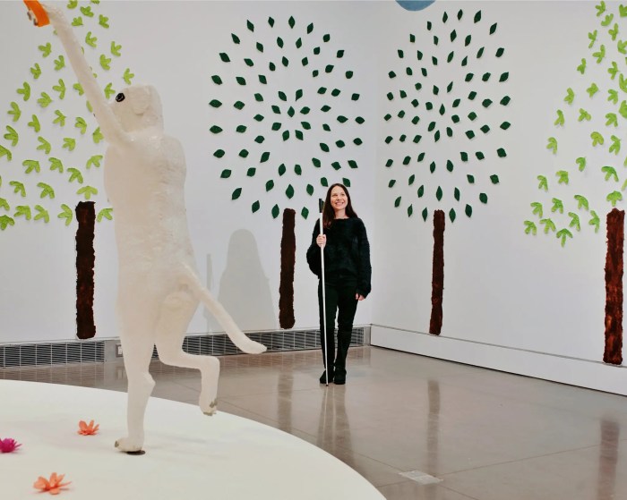 Artist Emilie Gossiaux holds a white cane as she stands in the room with her exhibit. Two trees with papier-mâché leaves are behind her.