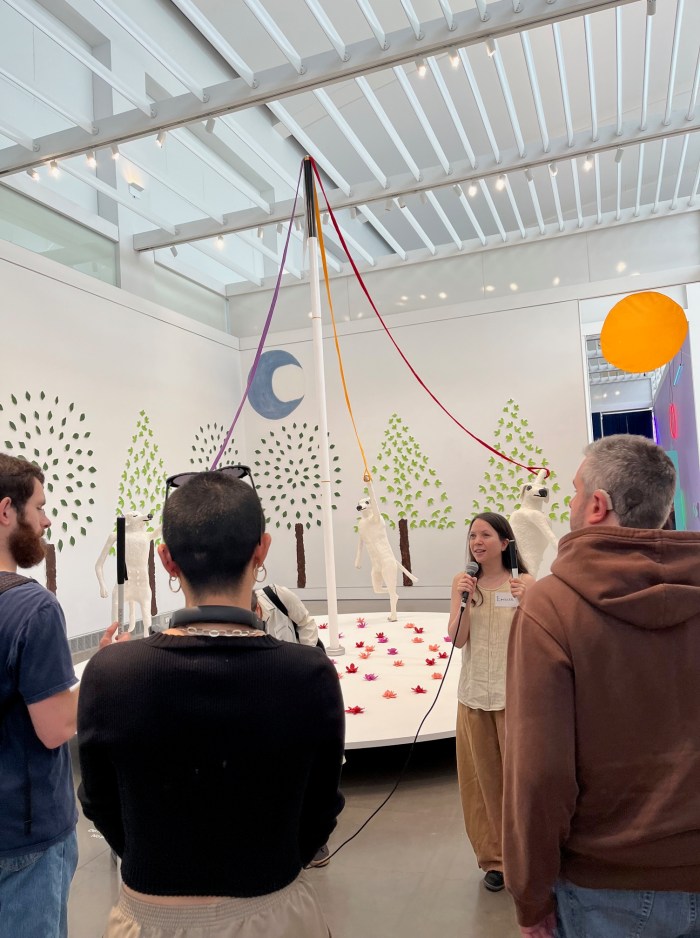 Artist Emilie Gossiaux stands in front of a 15-foot tall white cane and talks to visitors. Red and purple flowers are in the background.
