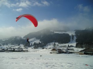 Person in a dark snowsuit with a red paraglider descends to land on a snowy field